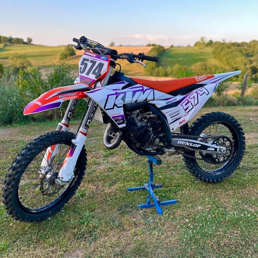 Motocross KTM avec kit déco HRL blanc, violet et orange, numéro 574, dans un champ